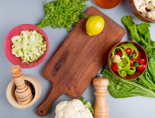 Top view of vegetables as bunch of coriander spinach sliced cabbage cauliflower sliced pepper with black pepper seeds and lemon on cutting board on blue background