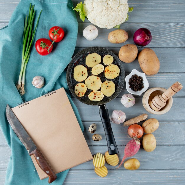 Top view of vegetables as cauliflower radish onion tomato and knife garlic crusher with pan of potato slices on wooden background