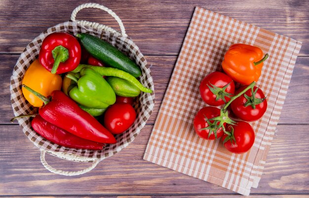 Top view of vegetables as cucumber pepper tomato in basket with tomatoes and pepper on plaid cloth and wooden surface