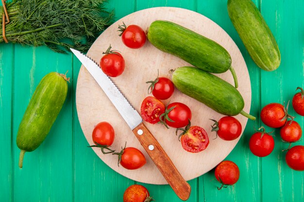 Top view of vegetables as cucumber and tomato with knife on cutting board and bunch of dill on green surface