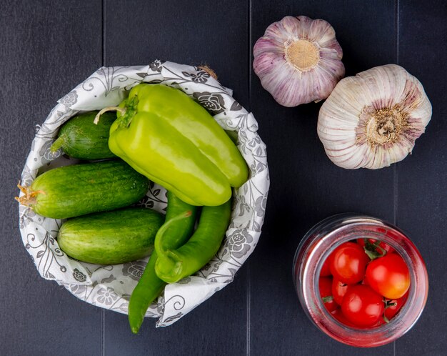 Top view of vegetables as cucumbers in basket and tomatoes in jar with garlic bulbs on black surface