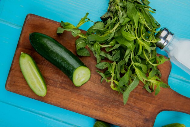 Top view of vegetables as cut cucumber and mint on cutting board with salt on blue surface