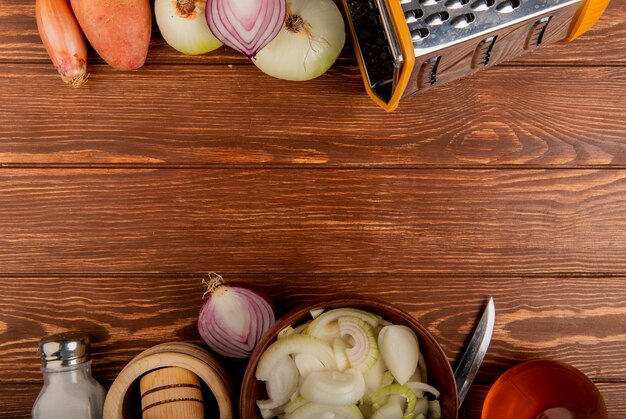 Top view of vegetables as different types of whole cut and sliced onions potato with salt butter knife and grater on wooden background with copy space