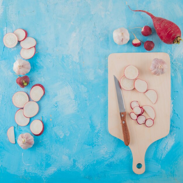 Top view of vegetables as garlic and radish with knife on cutting board on blue background with copy space