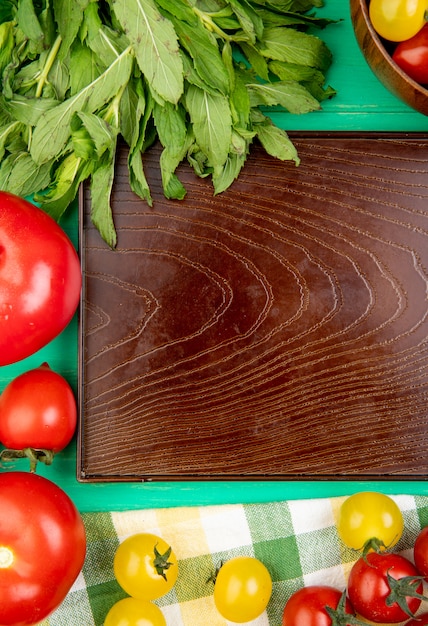 Top view of vegetables as green mint leaves tomatoes around empty tray on green surface