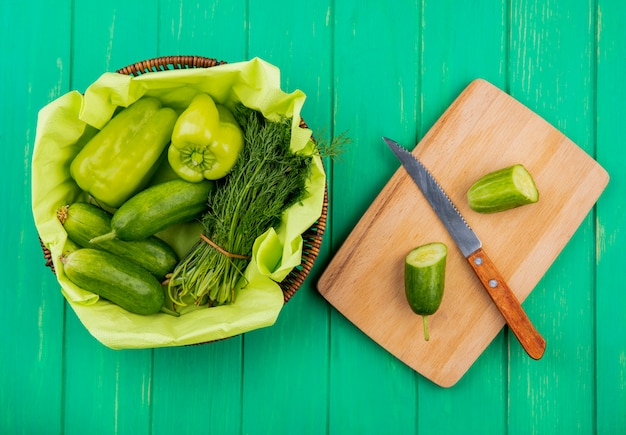 Top view of vegetables as pepper cucumber dill in basket with cut cucumber and knife on cutting board on green surface