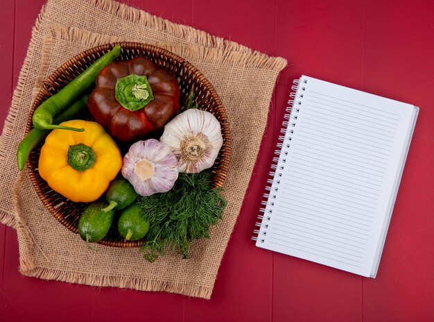 Top view of vegetables as pepper cucumber garlic dill in basket on sackcloth and note pad on red surface