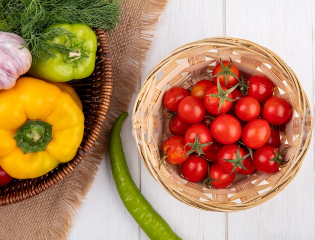 Top view of vegetables as pepper garlic dill in basket on sackcloth with basket of tomatoes on wooden surface