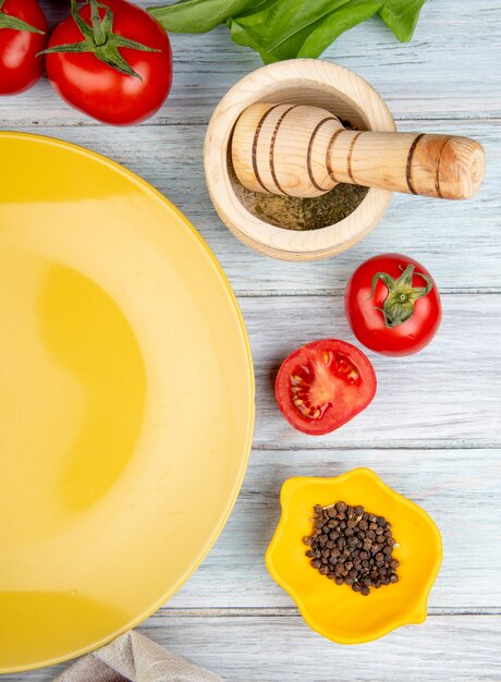 Top view of vegetables as tomato green mint leaves with black pepper seeds garlic crusher and empty plate on wood