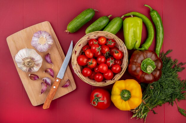 Top view of vegetables as tomatoes in basket cucumbers peppers dill and garlic with knife on cutting board on red surface