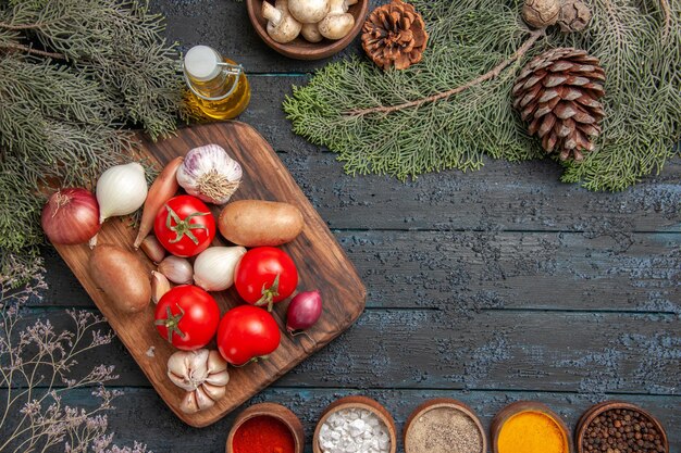 Top view vegetables and branches cutting board and vegetables on it between colorful spices and oil bowl of white mushrooms and spruce branches