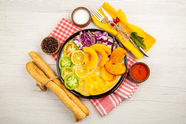 Top view vegetables and fruits cut lemons cut green tomatoes cut bell peppers cut persimmons on black plate red pepper powder sea salt black pepper in small bowls bread knife and fork on yellow napkin