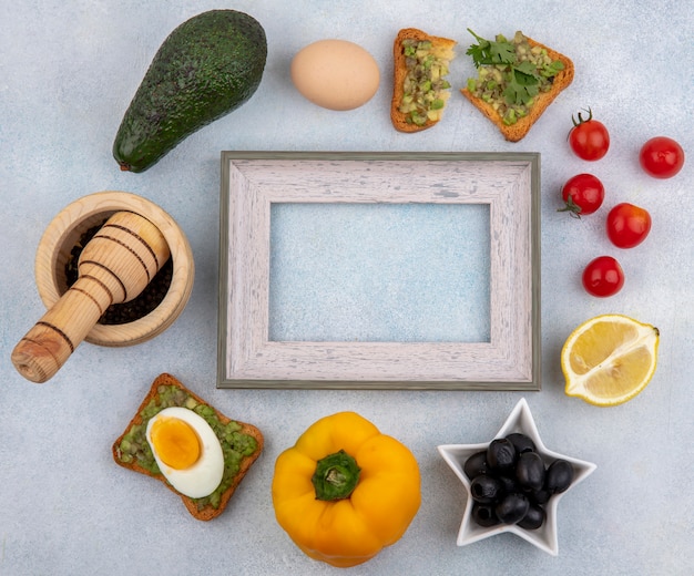 Top view of vegetables such as avocado lemon black olives yellow bell pepper tomatoes and a bread slice with avocado pulp on white surface