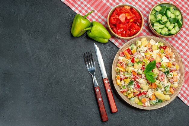 Top view of veggie salad in bowl with red napkin under it and cutleries with vegetables on side and place for text on dark grey background