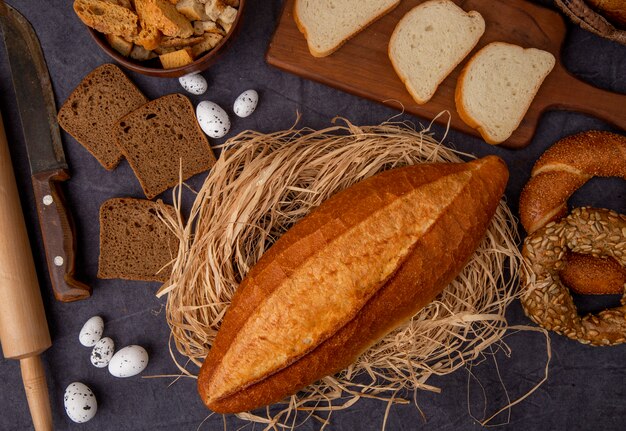 Top view of vietnamese baguette on straw with eggs knife bagels white and rye breads on maroon background