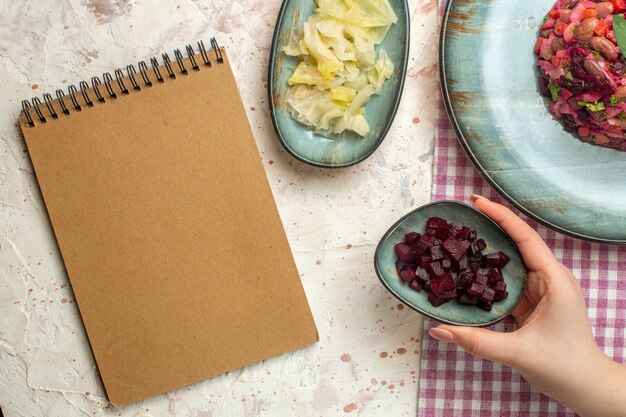 Top view vinaigrette salad on cyan oval plate on white and purple checkered tablecloth cut beet bowl in female hand notepad on light grey table