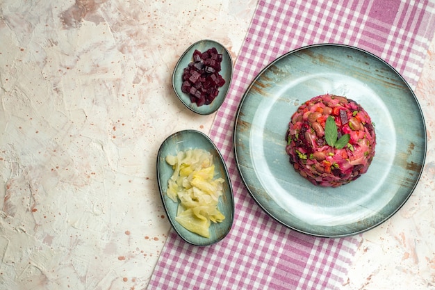 Top view vinaigrette salad on oval plate cut beet and other stuff in bowls on purple white checkered tablecloth on light grey table with free space