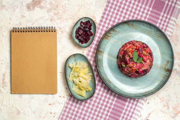 Top view vinaigrette salad on oval plate cut beet and other stuff in bowls on purple and white checkered tablecloth notebook on light grey table