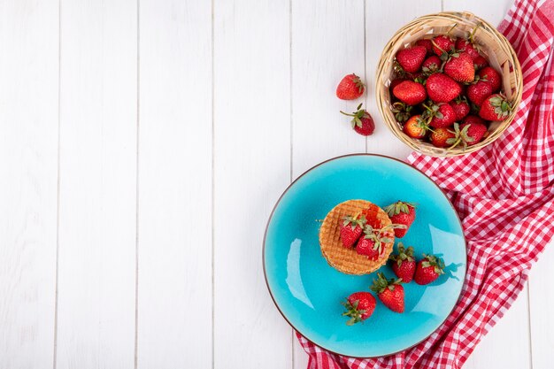 Top view of waffle biscuits and strawberries in plate and in basket on plaid cloth and wooden surface