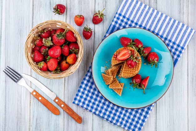 Top view of waffle biscuits and strawberries in plate and in basket with fork and knife on plaid cloth and wooden surface