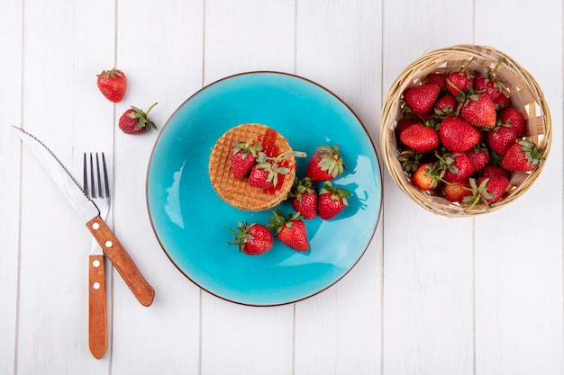 Top view of waffle biscuits and strawberries in plate and in basket with fork and knife on wooden surface