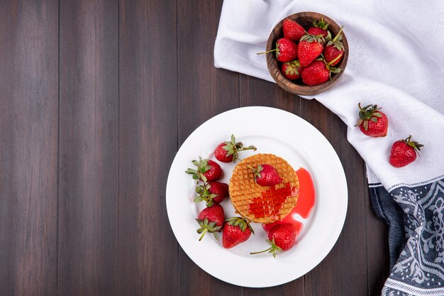 Top view of waffle biscuits and strawberries in plate and bowl of strawberries on cloth on wooden surface