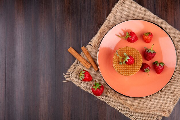 Top view of waffle biscuits and strawberries in plate with cinnamon on wooden surface