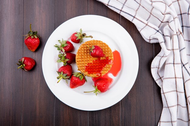 Top view of waffle biscuits and strawberries in plate with plaid cloth on wooden surface