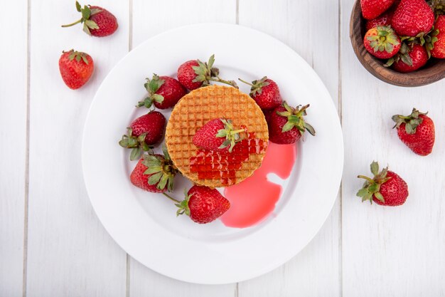 Top view of waffle biscuits with strawberries in plate and in bowl on wooden surface