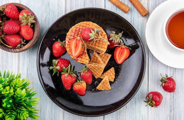 Top view of waffle biscuits with strawberries in plate and cinnamon cup of tea on wooden surface