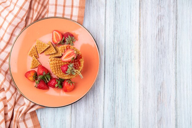 Top view of waffle biscuits with strawberries in plate on plaid cloth and wooden surface