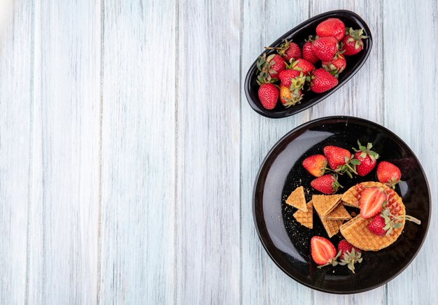 Top view of waffle biscuits with strawberries in plates on wooden surface