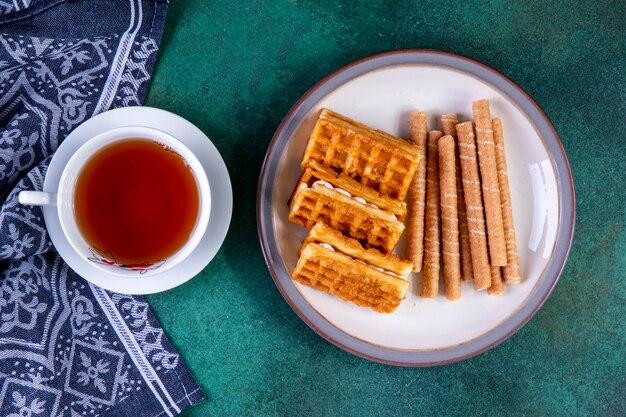 Top view waffles and sweet rolls on plate with a cup of tea on green