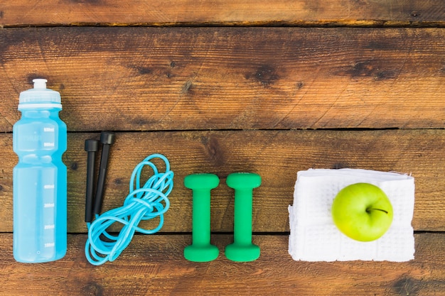 Free photo top view of water bottle; kipping rope; dumbbells; napkin and green apple on wooden background