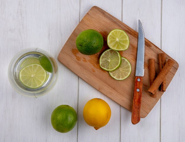 Top view water in a glass with lime and lemon on a board with a knife and cinnamon on a white table