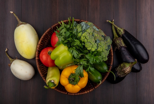 Top view white and black eggplant with broccoli  bell peppers  cucumbers and tomatoes in basket on wooden background