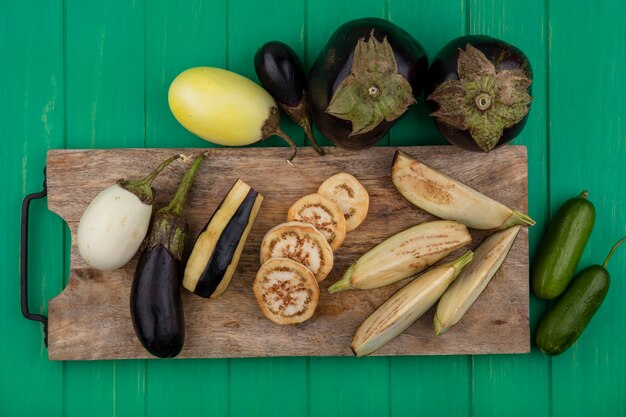 Top view white and black eggplant with slices on a cutting board with cucumbers on a green background