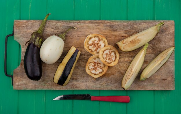 Top view white and black eggplant with slices on a cutting board with a knife on a green background