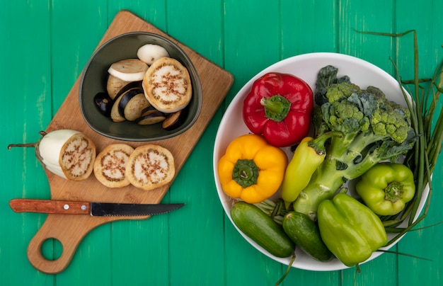 Top view white and black sliced eggplant with a knife on a cutting board with broccoli  cucumbers  green onions and peppers in a plate on a green background