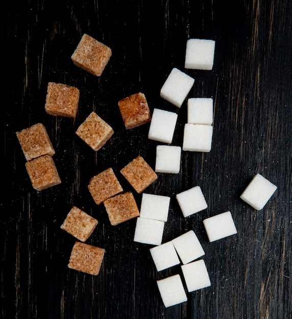 Вид сверху кубов белого и коричневого сахара, разбросанных на темном деревянном фоне