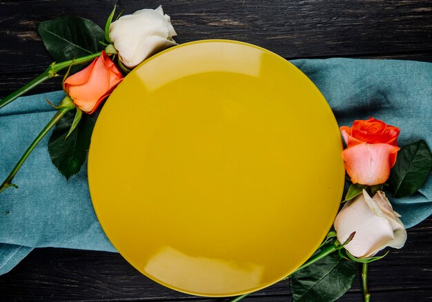 Top view of white and coral color roses arranged around a yellow plate on blue fabric on dark wooden background