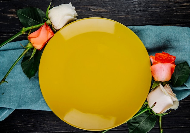 Top view of white and coral color roses arranged around a yellow plate on blue fabric on dark wooden background