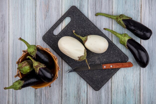 Free photo top view of white eggplants and knife on cutting board with black nadia ones in basket and on wooden background