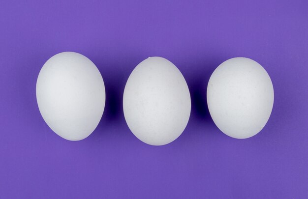 Top view of white fresh chicken eggs arranged in a line on a violet background