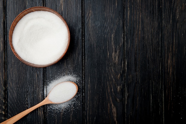 Top view of white granulated sugar in a wooden bowl and in a wooden spoon on black rustic background with copy space