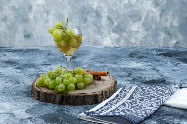 Top view white grapes, cinnamon, glass of whisky on wooden board with kitchen towel on dark and light blue marble background. horizontal