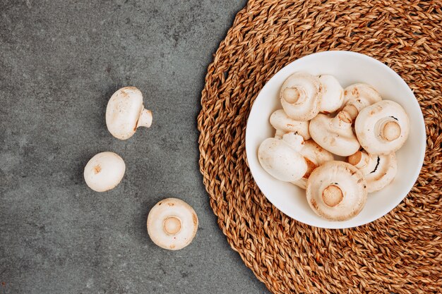 Top view white mushrooms in bowl on rattan trivet and gray textured background. horizontal