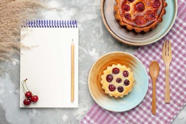 Top view white notepad with little cake and raspberry cake on the light desk fruit berry bake cake pie