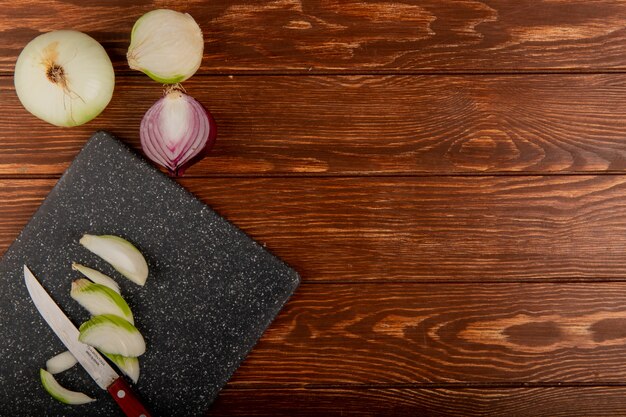 Top view of white onion slices and knife on cutting board with whole ones and half cut red onion on wooden background with copy space