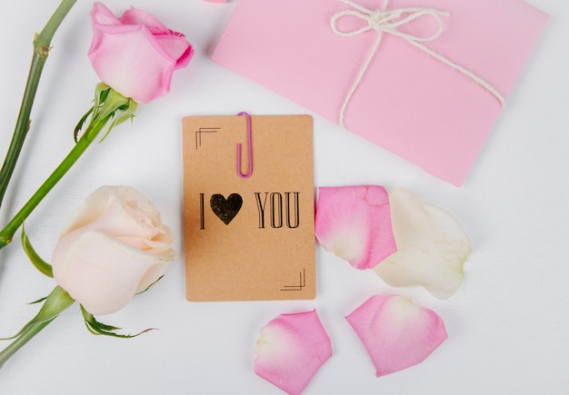 Вид сверху белых и розовых цветных роз с конвертом, перевязанным веревкой и небольшой открыткой со скрепкой и лепестками роз на белом фоне